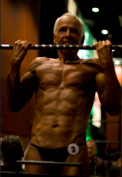 A man in late adulthood is doing pull-ups at a body-building championship. 