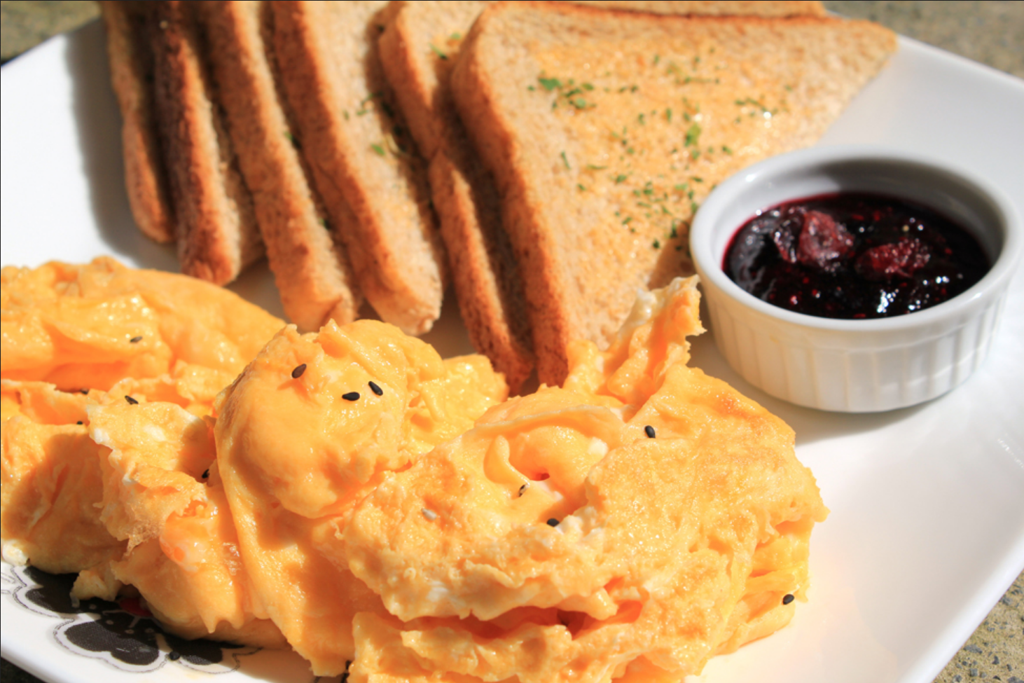 A plate with a large pile of eggs and six slices of toast. There is a small dish of red jam near the toast on the plate.