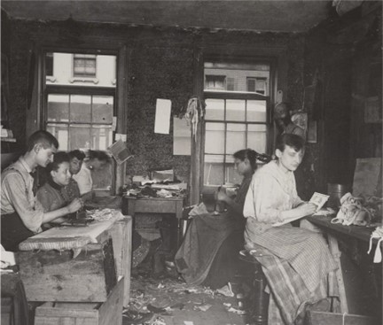 Photograph shows men and women sewing and ironing in tenement factory, probably in New York City.